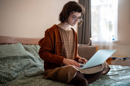 Photo for Busy thoughtful teenage girl sitting on bed with laptop computer, thinking, reading text document, doing homework while studying online, working as freelancer, blogger, writing book in home atmosphere - Royalty Free Image