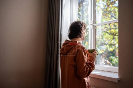 Photo for Lonely teenager standing near window, looking at sunny summer weather with sadness, sorrow, spending time alone at home. Unsociable teen girl having no friends. Youthful crisis, communication problems - Royalty Free Image