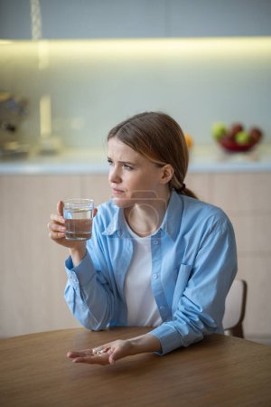 Photo for Sick middle-aged female sitting at table with pills, painkillers, antibiotics on palm, hold glass of water, looking at window, going to take treatment. Depressed girl suffering from headache, pain - Royalty Free Image