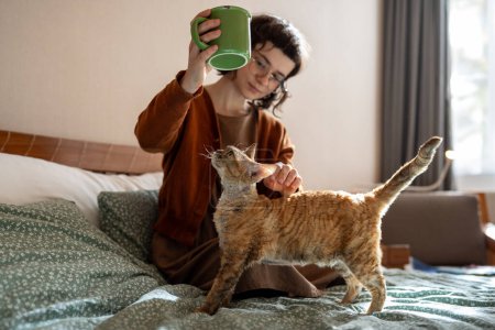 Foto de Chica adolescente sentada en la cama, sosteniendo una taza de té, yendo a beber mientras el amado gato Devon Rex quiere jugar, requiere acariciar, acariciar. Animal doméstico que proporciona apoyo emocional, comodidad al dueño de la mascota - Imagen libre de derechos