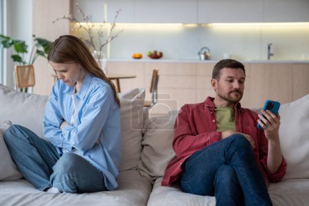 Photo for Jealous wife sad about husband messages in phone sitting on couch at home. Spouses ignoring each other. Upset woman turn out from man who looking at smartphone. Betrayal infidelity mistrust concept. - Royalty Free Image