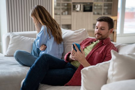 Photo for Woman crying turning out of man sitting on couch at home. Husband ignoring wifes emotions looking at smartphone screen. Couple spouses misunderstanding in relationships, jealous mistrust concept. - Royalty Free Image