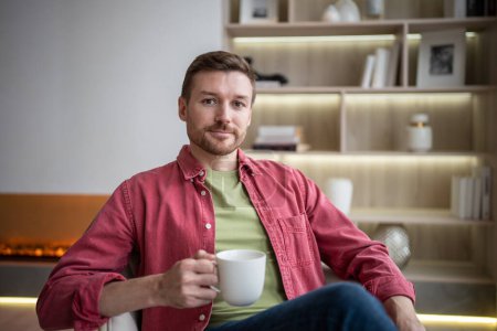Photo for Positive carefree middle-aged bearded man resting on chair at apartment. Cheerful male sitting at home, holding cup, drinking tea, looking at camera friendly smiling. Wellbeing, slow living concept - Royalty Free Image