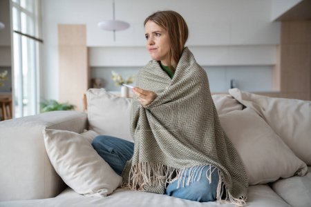 Photo for Sick woman checking temperature with thermometer sitting on sofa at home wrapped in plaid. Sad frustrated young girl suffering from seasonal flu or cold. Worry about feeling unhealthy with influenza. - Royalty Free Image