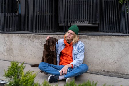 Photo for Upset depressed millennial guy sitting on sidewalk with dog, having housing problems. Lonely young man on city street thinking about life problems. Emotional support animal concept - Royalty Free Image