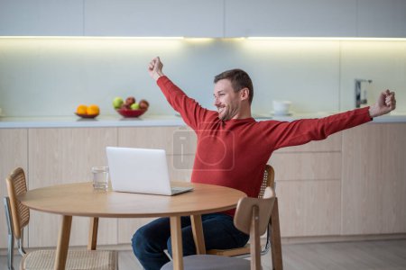 Téléchargez les photos : Homme freelance heureux étirant les bras souriant travail de fin assis sur la cuisine avec ordinateur portable. Le gars qui se repose après avoir travaillé sur ordinateur à la maison. Freelance, travail à distance, travail, projet de finition. - en image libre de droit