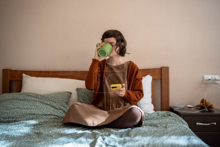 Photo for Introverted teenage girl sitting on bed, drinking tea, spending pastime at home looking photos in social networks, reading online news. Lazy girl addicted to smartphone, doing nothing at home - Royalty Free Image
