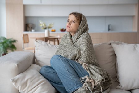 Photo for Sad tired woman sits on couch at home wrapped in plaid in living room. Blue mood of depressed girl boring. Nervous female feeling frustration hiding from society in loneliness solitude. Life problems. - Royalty Free Image