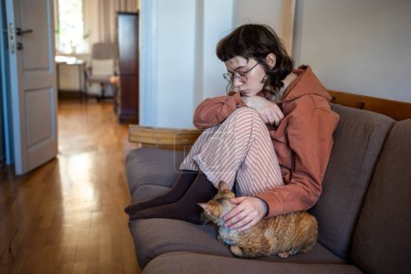 Depressed exhausted teen girl in glasses pyjamas petting cat sitting on couch at home in closed pose. Teenage age crisis, emotional burnout depression, psychological or life problems troubles concept.