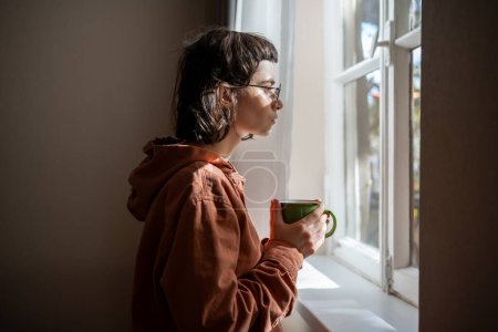 Téléchargez les photos : Adolescent triste solitaire avec une tasse de thé regardant une fenêtre, couvant. Adolescente insociable passant le week-end à la maison en raison de l'amitié, des difficultés sociales, des problèmes de communication, de malentendu - en image libre de droit