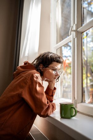 Photo for Nerd teen girl in glasses drinking tea looking at window boring at home. Sad upset young woman feeling apathy, age teenage crisis. Mental disorder, anhedonia, stress, having life troubles problems. - Royalty Free Image