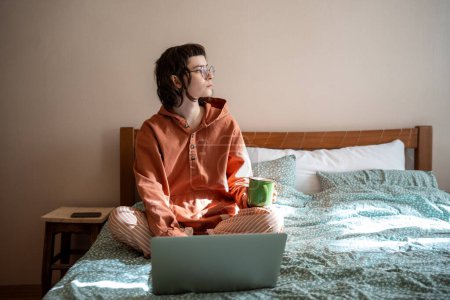 Photo for Pensive teenage girl sitting on bed with cup of tea, looking at window, thinking, having tea break while doing homework, studying on laptop computer. Home schooling, distant online education concept - Royalty Free Image