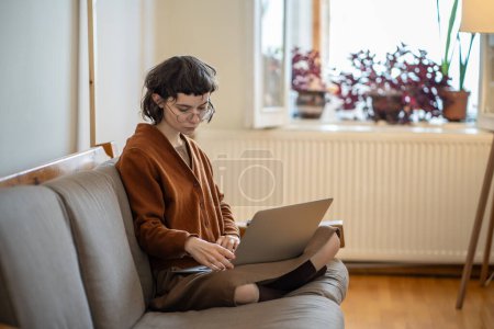 Téléchargez les photos : Étudiante adolescente concentrée étudiant sur ordinateur portable assis sur le canapé dans la pose de lotus à la maison. Femme occupée dans des lunettes travaille sur un projet éducatif, fait des devoirs regarde l'écran d'ordinateur. Concept d'éducation. - en image libre de droit