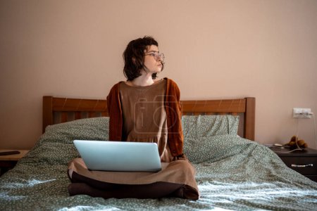 Photo for Pensive teenager sitting on bed with laptop computer on knees, resting after long freelance work, looking at window, thinking. Teen girl looks like online tutor, writer, journalist, blogger, student - Royalty Free Image