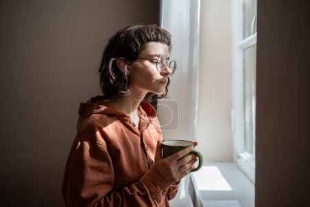 Photo for Teen girl nerd in glasses looking at window drinking cup of tea at home. Serious young woman resting contemplating street, watching people cars. Weekend, of introvert, leisure in loneliness concept. - Royalty Free Image