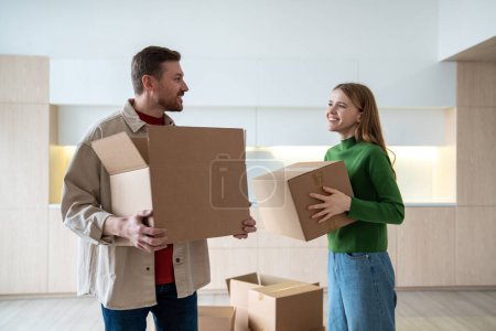 Photo for Happy new homeowners tenants renters pleased man and woman carrying boxes to room. Family couple looking eye to eye standing on kitchen while moving to apartment. Relocation, family mortgage concept. - Royalty Free Image
