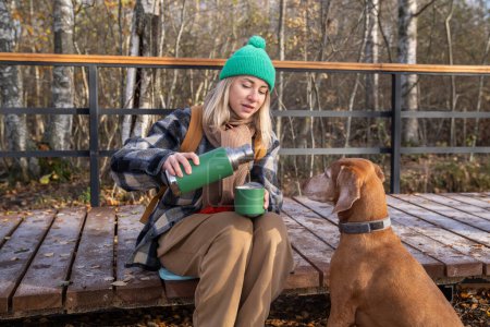 Foto de Mujer sirviendo té de termos sentado en el camino de madera en el parque de otoño viajando con el perro. Propietaria de mascotas de mediana edad que descansa en caminatas por el bosque disfrutando de la naturaleza. Viajes, turismo, ansia de viajar. - Imagen libre de derechos