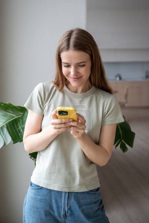 Photo for Contented relaxed woman reading messages in mobile phone at home near window. Smiling girl chatting in smartphone with friends. Positive young female enjoy relaxing in gadget. Social media addiction. - Royalty Free Image