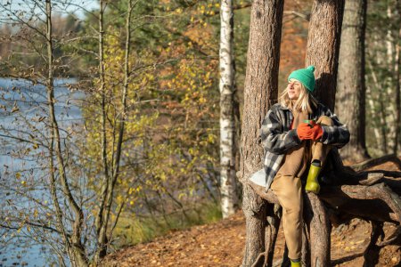 Photo for Carefree tourist woman hiker camper drinking hot tea from cup enjoying nature forest lake. Female admiring Scandinavian woodland, heating with beverage in autumn park. Love of nature, mental health - Royalty Free Image