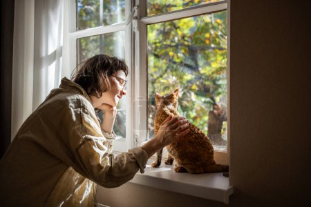Foto de Gato de raza Devon Rex sentado en el alféizar de la ventana, calentándose a la luz del sol. Adolescente cariñosa mirando al gatito con amor, ternura. Adolescente acariciando, acariciando gatito ronroneando de placer. Amor incondicional a las mascotas - Imagen libre de derechos