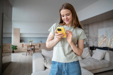 Photo for Pleasantly smiling cheerful positive middle-aged European woman standing in modern apartment, holding mobile phone, reading, writing message, chatting, scrolling photos online in social network - Royalty Free Image