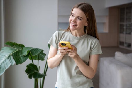 Photo for Jolly cheery friendly smiling positive middle-aged European woman standing near window, holding mobile phone, messaging, typing, looking into street, waiting for guests, online shop delivery courier - Royalty Free Image