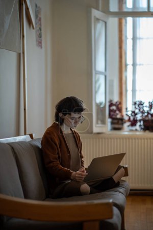 Photo for Focused teen girl working on laptop, using social media sitting on couch at home. Young woman in glasses looking at computer screen messaging online reading typing messages. Digital entertainment. - Royalty Free Image