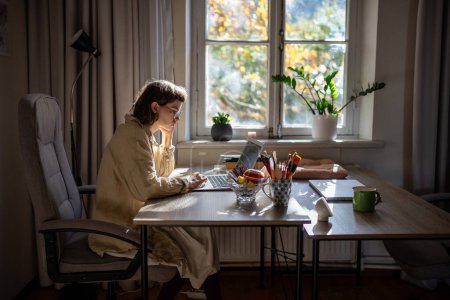 Téléchargez les photos : Adolescente réfléchie assise à table à la maison. Adolescent diligent concentré lisant le texte de l'écran d'ordinateur portable, pensant, tout en travaillant comme pigiste, étudiant en ligne sur l'éducation à distance - en image libre de droit