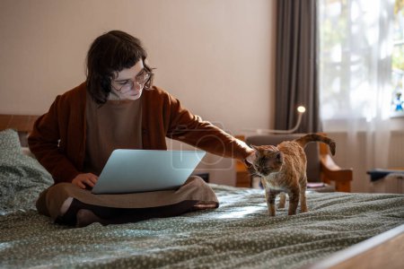 Téléchargez les photos : Adolescente étudiante étudiant sur ordinateur portable assis dans le lit à la maison caressant chat. Adolescente brune nerd dans des lunettes matériel d'apprentissage. Je m'occupe du chat domestique. Éducation en ligne, faire ses devoirs. - en image libre de droit