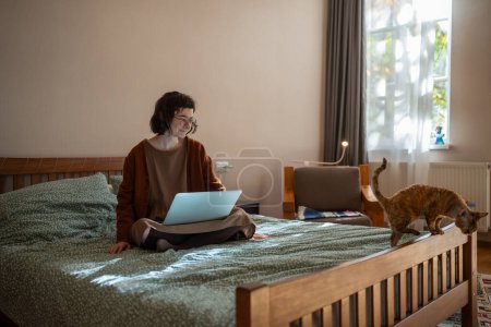 Photo for Woman freelancer working on laptop sitting in bed looking at pet cat at home. Girl in glasses having problems with self-organization of distant work, distracted from work. Freelance online job concept - Royalty Free Image
