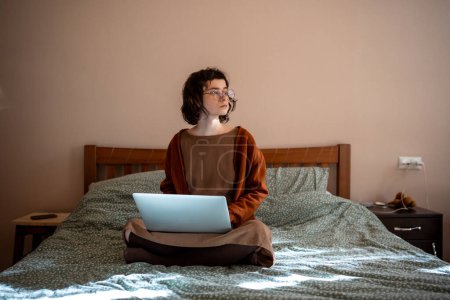 Photo for Teen girl student studying on laptop sitting in bed at home looking at window take a break. Nerd teenager in glasses learning materials on computer. Distant, remote education, doing homework concept. - Royalty Free Image