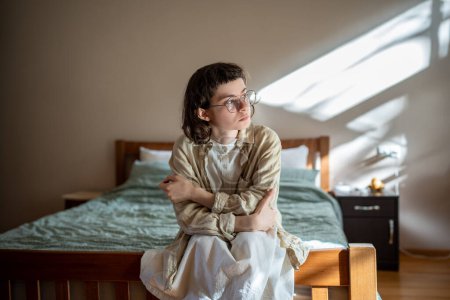 Photo for Reserved melancholic unemotional teenager sitting on bed, indifferently looking at window, feeling solitude, sadness, absence of motivation, procrastination. Adolescence, puberty, awkward age problems - Royalty Free Image