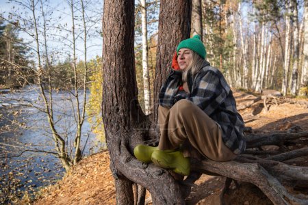 Photo for Pensive smiling midle-aged woman sitting on pine tree root, enjoying sunny warm autumn day, thinking of life, dreaming, listening to silence. Recreation, relaxation in nature, pleasant time spending - Royalty Free Image