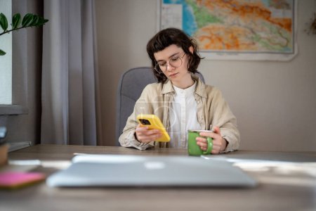 Photo for Serious teenage girl having tea break after freelance work, studying on distant education. Pensive teen sitting at table with cup of hot beverage, holding smartphone, scrolling web pages in internet - Royalty Free Image
