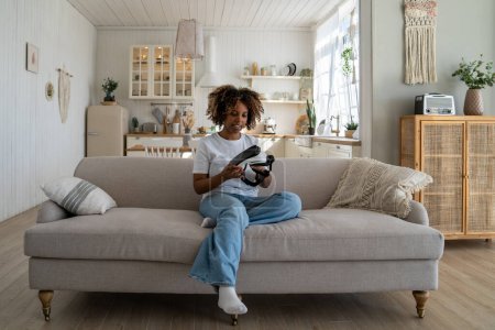 Téléchargez les photos : Fille afro-américaine millénaire avec les cheveux bouclés assis sur le canapé tester un nouveau casque VR, en utilisant le casque de réalité virtuelle pour voir des photos et des vidéos en 3D. Jeune femme examinant des lunettes AR à la maison - en image libre de droit