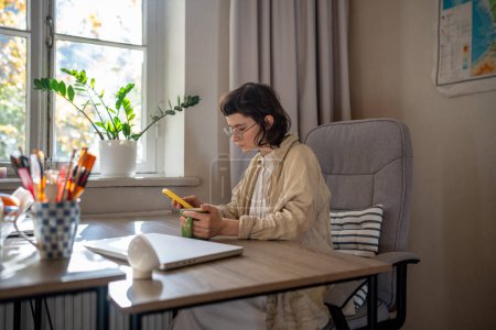 Photo for Concentrated teenage girl sitting at table at home, holding smartphone, reading news, information, messaging. Laptop computer lying on table. Diligent teen looks like freelancer, student, tutor - Royalty Free Image