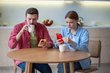 Photo for Pensive couple obsessed with smartphones sitting in kitchen, drinking tea, ignoring each other. Concentrated on mobile phones man and woman checking social networks, scrolling web pages, photos, news - Royalty Free Image
