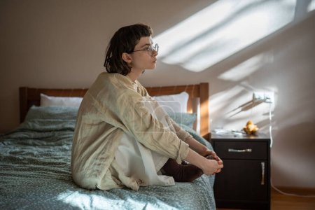 Photo for Introverted unemotional teenager sitting on bed, indifferently looking at window, feeling loss of interest to life, absence of motivation, procrastination. Adolescence, puberty, awkward age problems - Royalty Free Image