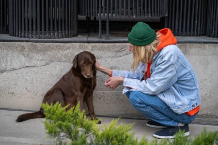 Photo for Long-haired stylish hipster guy sitting on haunches, stroking, caressing homeless stray dog in city street. Kind-hearted male looking at animal with love. Kindness, compassion, mercifulness to animals - Royalty Free Image