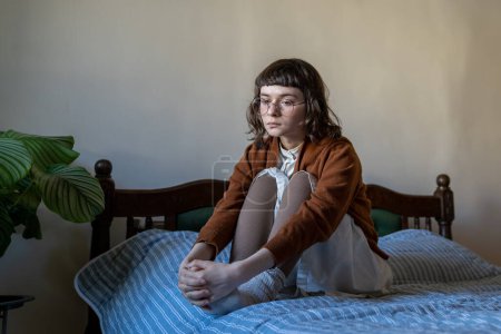 Unemotional introverted teenager with empty glance sitting on bed alone at home. Unhappy sed girl in depression, solitude, having no motivation, interest to life. Adolescence, puberty period problems