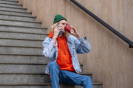 Téléchargez les photos : Homme mince élégant portant un sweat à capuche coloré à la mode et un jean assis sur un escalier, parlant sur smartphone, buvant du café. Homme aux cheveux longs ressemble à touriste, modèle, étudiant, IT freelance, blogueur - en image libre de droit