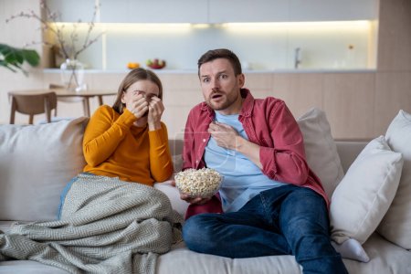 Scared wife and husband watch thriller eat popcorn sitting on home couch. Afraid family couple enjoy horror movie series. Fearful woman closing face with hands while seeing movie terrible episode