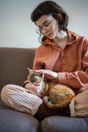 Caring pet owner stroking petting cat sitting on couch at home enjoying communication. Teen girl in pyjama cat lover spending time with pet. Cat lying on loving woman legs closing eyes from pleasure.