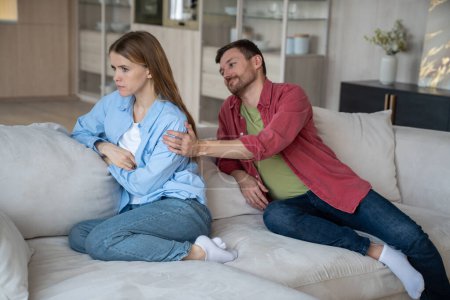 Photo for Offended woman with indifferent apathetical glance, sitting on sofa, turning back to husband, unwilling to speak. Caring man calming down irritated angry female. Conflict, crisis in family relations. - Royalty Free Image