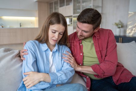 Tender caring husband sitting on sofa, calming down upset depressed wife in grief, sorrow. Loving man embracing sad female. Emotional support, consolation in difficult situations in family relations