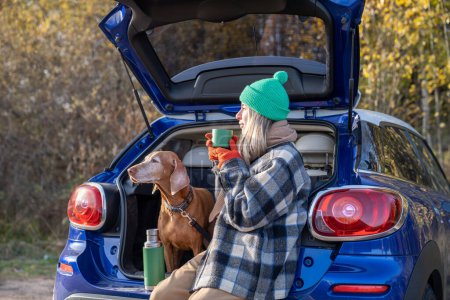 Photo for Woman pet owner enjoying drinking tea sitting in car trunk with dog admiring nature in forest on weekend. Autumn outdoor activity Scandinavian vacation travel tourism road trip with magyar vizsla dog. - Royalty Free Image
