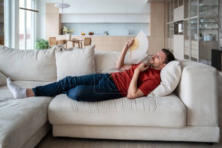 Photo for Overheated perspiring man lying on sofa, waving with hand fan, suffering from high temperature in room because of broken conditioner, absence of ventilator, air blower. Male feeling unwell, dehydrated - Royalty Free Image