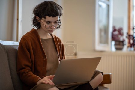 Téléchargez les photos : Femme freelance focalisée travaillant sur ordinateur portable, étudiant en ligne, assise sur le canapé avec ordinateur sur les genoux, pensant, lisant le texte de l'écran, travaille sur un nouveau projet. Travail confortable dans l'atmosphère de la maison - en image libre de droit