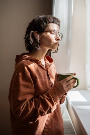 Photo for Pensive introvert woman in glasses looking at window drinking cup of tea at home. Girl suffering from mental illness looks outside, watching people, cars, thinking about life problems. Loneliness. - Royalty Free Image