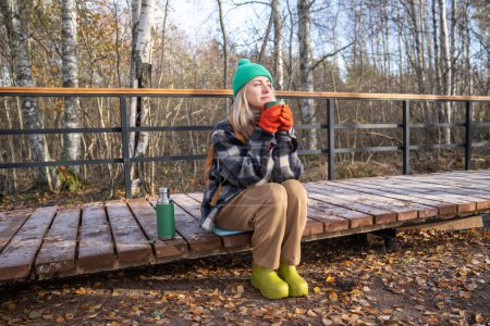 Téléchargez les photos : Femme détendue randonneur chauffage boire du thé chaud assis sur le chemin en bois dans le parc. Calme femelle d'âge moyen reposant dans la forêt profitant de la nature sur un sentier en bois. Voyages, tourisme, slow life, wanderlust - en image libre de droit